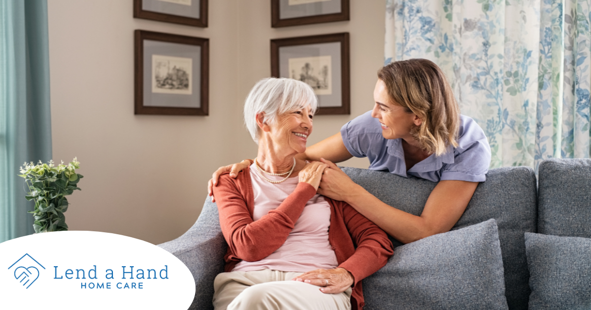 Smiling caregiver with hand over the shoulder of a happy elderly woman sitting on the couch.