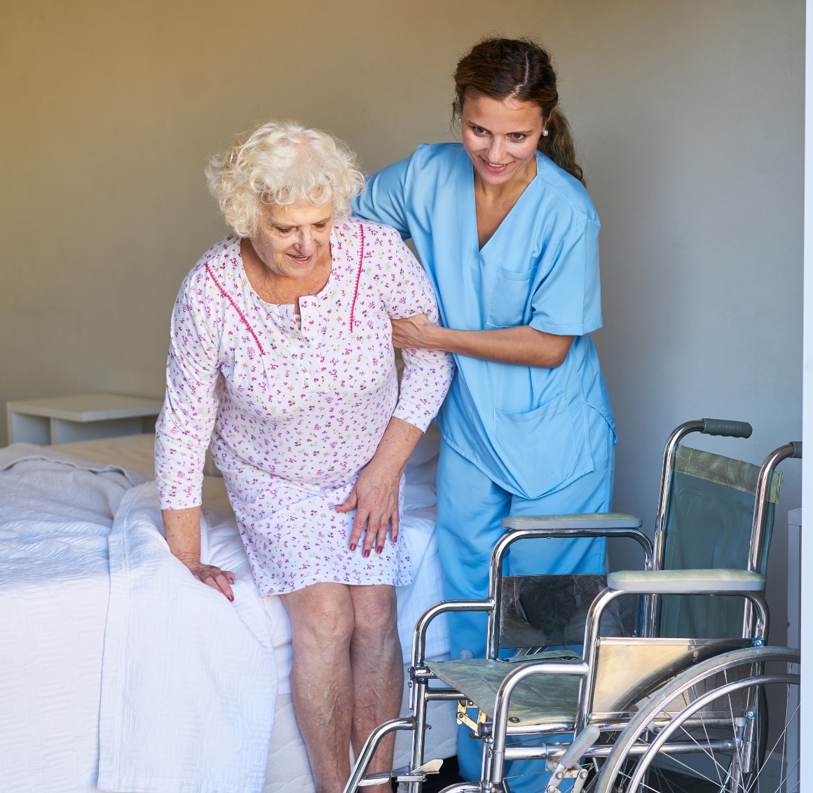 companion care in sparks – 1 A young female caregiver in light blue scrubs assists a senior woman as she carefully helps her sit in a wheelchair beside a bed in a cozy bedroom. The scene reflects gentle support and attentive care.